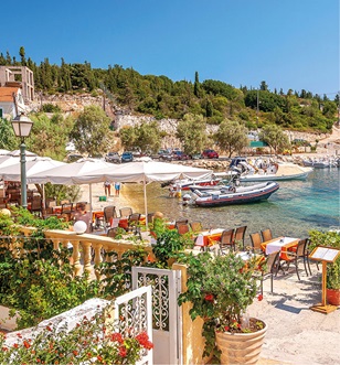 restaurant by the water with white walls, green plants with red flowers, tables outside and boats in the water