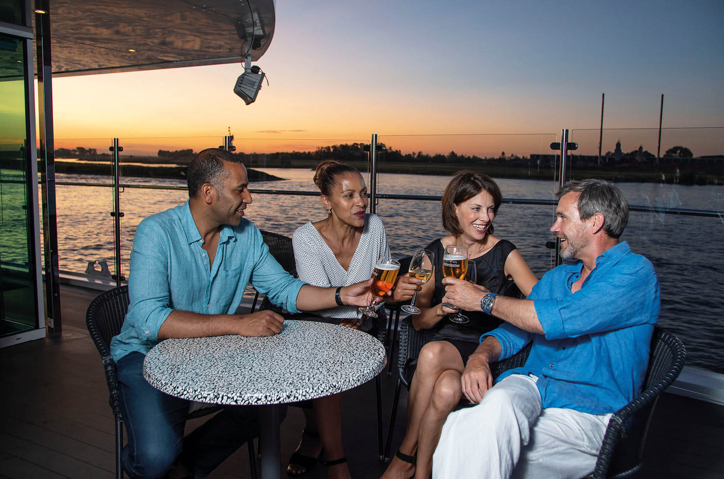 Four friends sitting outside on a cruise ship enjoying a drink as the sun sets behind them