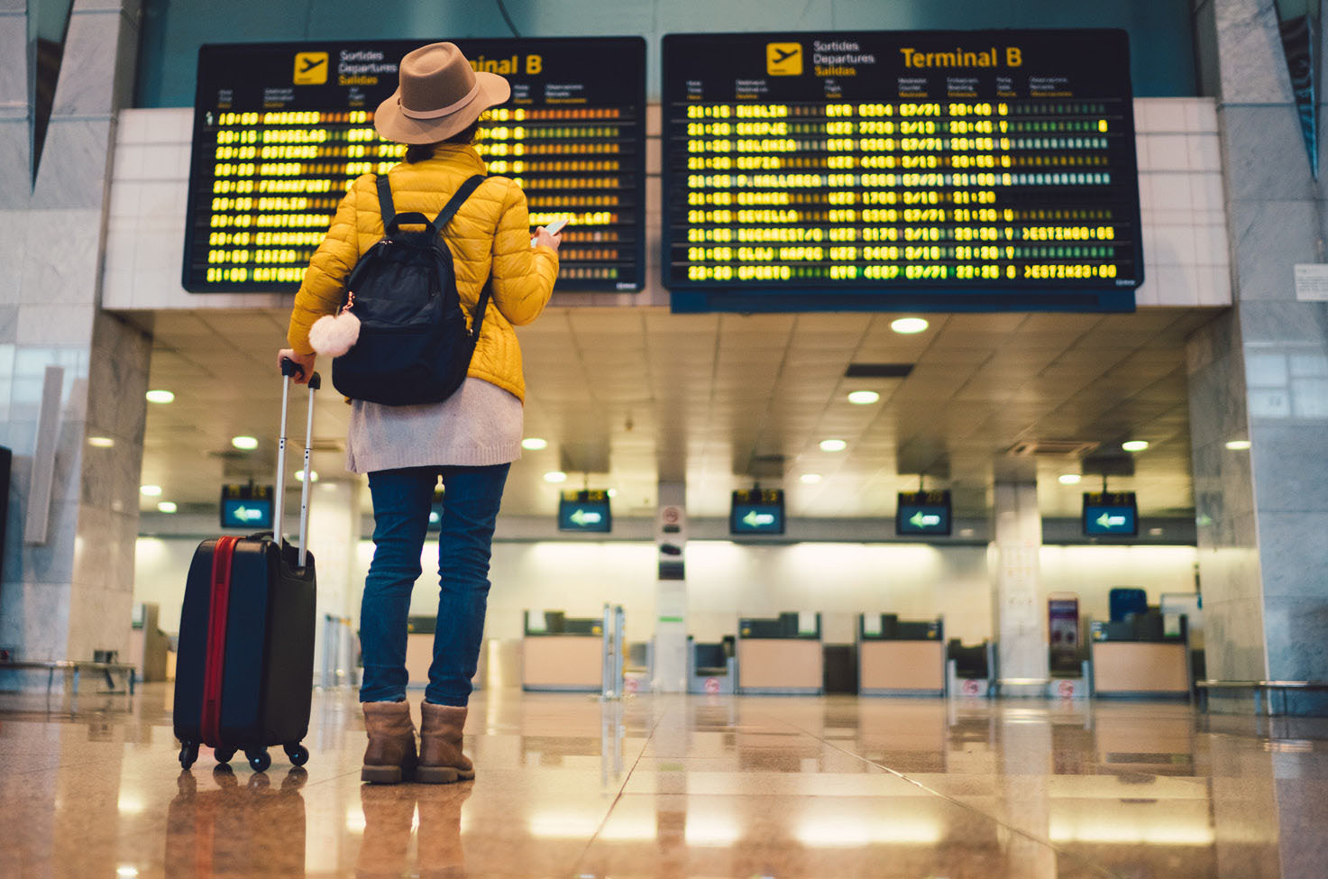Person wearing yellow jumper, with their bag and suitcase, checking flight departure times in an airport