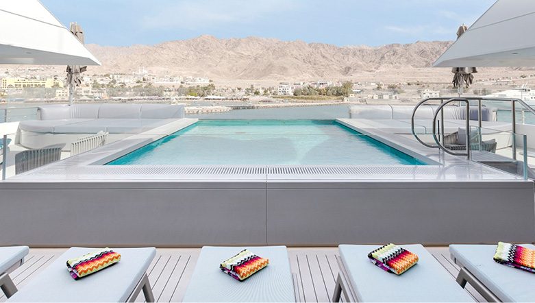 A large pool on the top deck of a yacht next to a row of sun loungers, with hills in the background